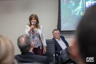Woman speaking into a microphone at a presentation. Man in background. People in the audience.