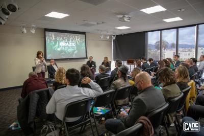 Panel discussion in a room with an audience. A woman speaks at the front, screen displays 