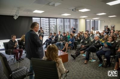 A man speaks to a group in an office setting; people are seated, listening.