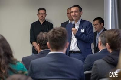 Man in blue suit speaks into microphone; audience seated.
