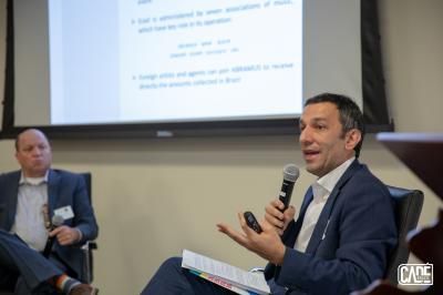 Two men at a panel. One speaks into a microphone, gesturing. The other watches. Presentation screen in the background.