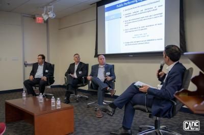 Panel discussion: Four men in suits; one speaks into a mic, others listen. Presentation screen visible.