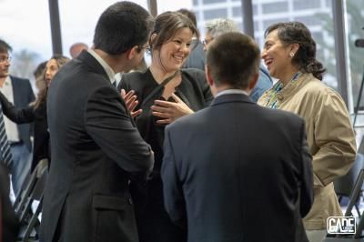 People in business attire chatting at an event, some smiling. Blurred background, likely an office.