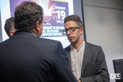 Man in glasses talking to two men at an event with a projector screen in the background.