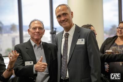 Two men smiling, posing together indoors; one gives thumbs up, both wearing suits, in a well-lit room.