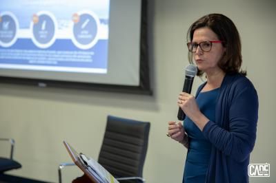 Woman speaking into a microphone at a conference; screen with diagrams in background.