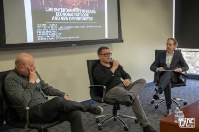 Three men seated for a panel discussion on live entertainment in Brazil. A projection screen is behind them.