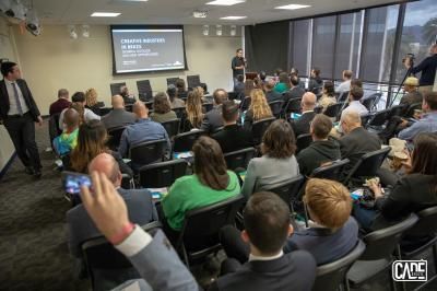Audience in a conference room, a speaker presenting on screen. People are seated, some taking photos.