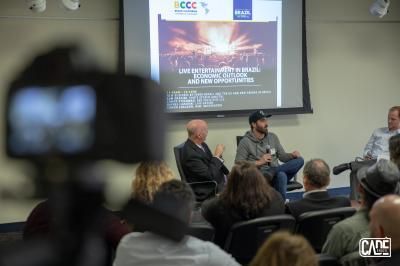 Panel discussion. Three men sit on stage, a screen with a presentation behind them. A camera records them from the audience.