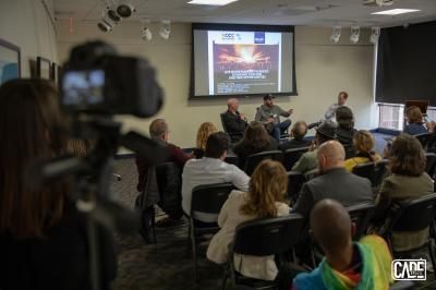 A panel discussion in a room, featuring speakers, audience, and a camera filming the event.
