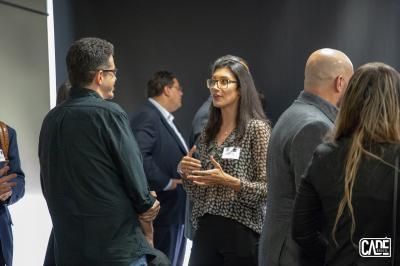 Woman in glasses speaks to a man at a networking event. Others in suits are nearby.