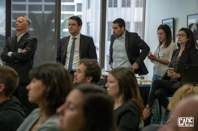 People in business attire in an office setting; some standing and looking forward, some seated, listening.
