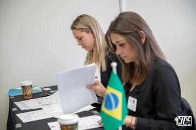 Two women reviewing documents at a table, Brazilian flag nearby.