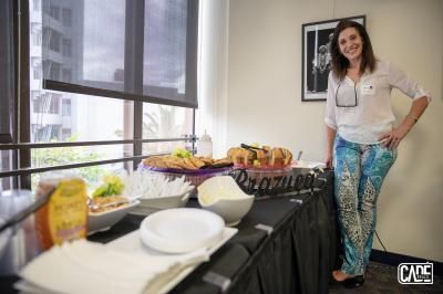 Woman standing near a table of food, smiling. She wears a white shirt, patterned pants. Sunlight from a window.