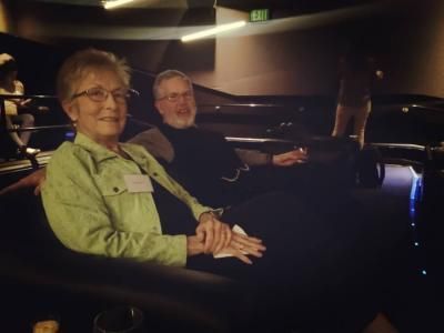 Elderly couple seated in a dark movie theater. Woman in green jacket smiles; man in black turtleneck looks on.