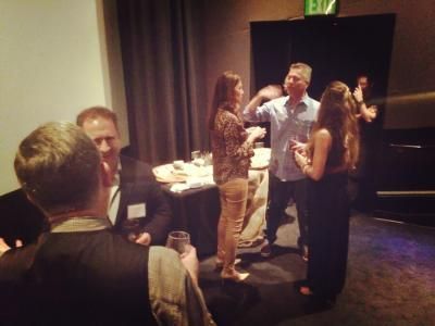 People socializing at an event, some holding drinks. Dimly lit room, table with food.