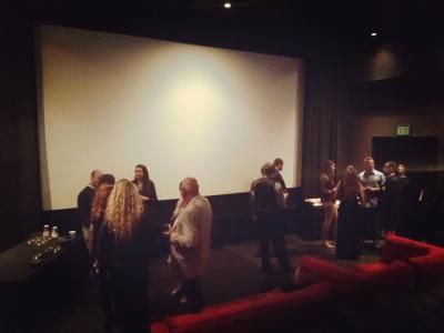 People mingling in a dimly lit movie theater before a screening, red seating visible, white screen, exit sign.