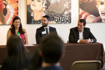 Three panelists seated at a table in front of an audience, smiling, discussing. Artwork hangs on the wall.