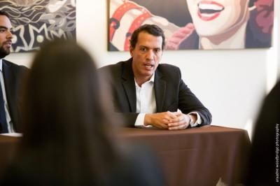 Man in a black suit speaks at a table, hands clasped, in a room with art on the walls.