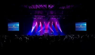Concert stage with purple and blue lights, band silhouetted, audience watching.