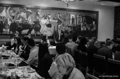 People seated at tables in a restaurant; a man in a suit speaks, mural in background.