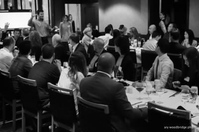 Black and white photo of people at tables, likely a banquet or reception.  A man is gesturing enthusiastically.