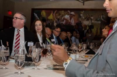 People seated at a formal dinner table, with a man in the foreground holding a card and others in the background.