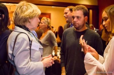 People talking at an event: blonde woman in a light suit, man in a green shirt, woman in white.