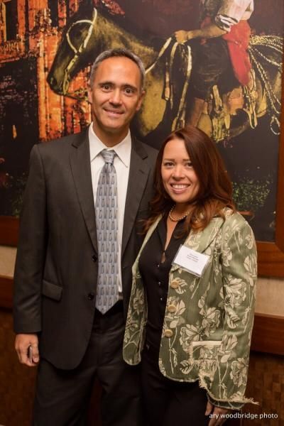 Man and woman standing together, smiling. Man in suit, tie, woman in patterned jacket. Background painting.