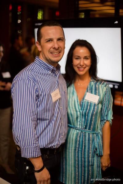 Man and woman smiling at an event, standing close together. Man in blue striped shirt, woman in blue patterned dress.