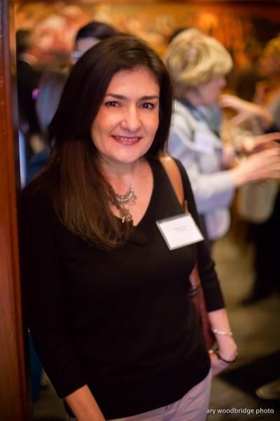 Woman with dark hair, wearing black top and name tag, smiling at an event, people in background.