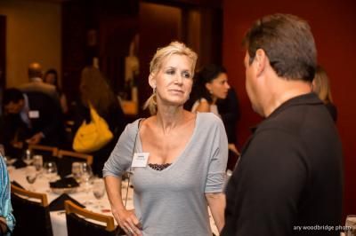 Woman in gray top, talking to a man in a social setting, near tables set for dining.