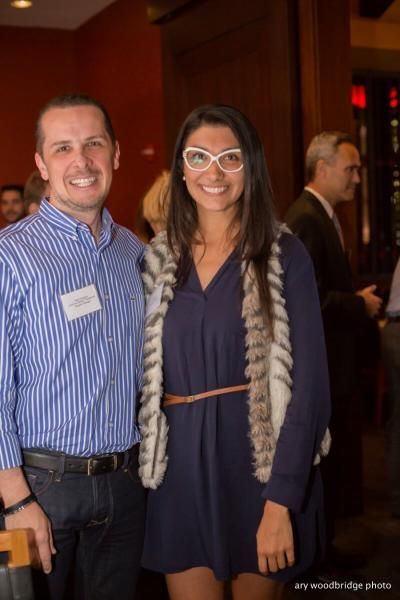 Man and woman smiling at a social event. Man in blue striped shirt, woman in blue dress and fur vest, glasses.