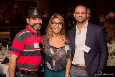 Three smiling people at an event: man in hat, woman in glasses, man in blue blazer.