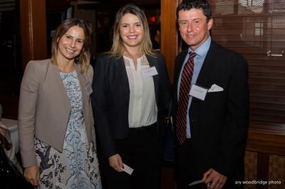 Three people in business attire at an event. Woman on the left smiles, two in the middle stand, all with nametags.