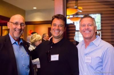 Three smiling men in an interior setting. One bald man in glasses, another with dark hair and a black shirt, and one in a blue striped shirt.