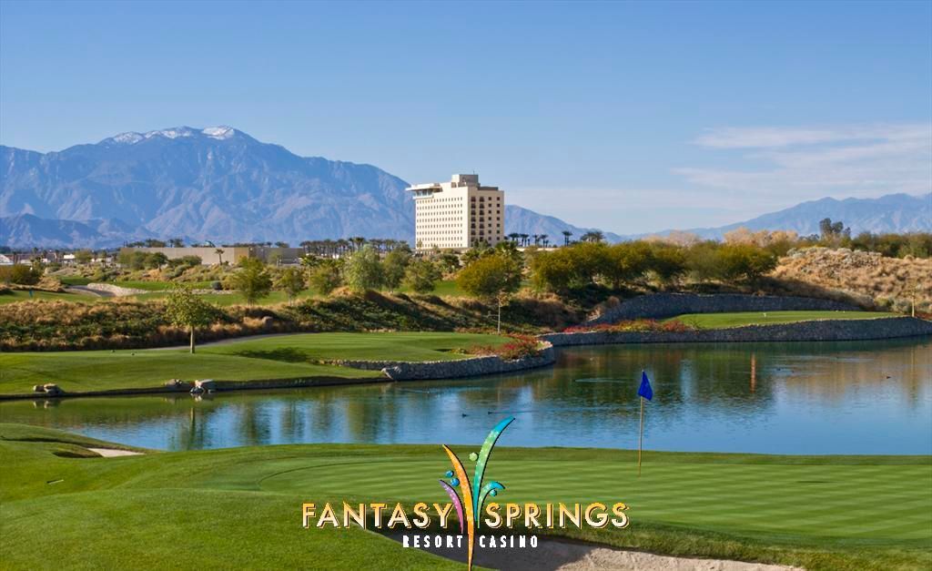 Fantasy Springs Resort golf course with lake, green, and hotel in the background, with mountains and a clear blue sky.