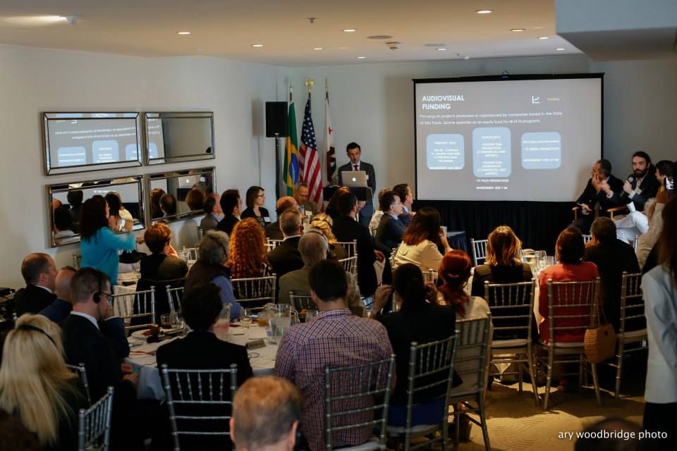 A presentation in a conference room. A speaker, audience, a projector displaying slides, and flags are visible.