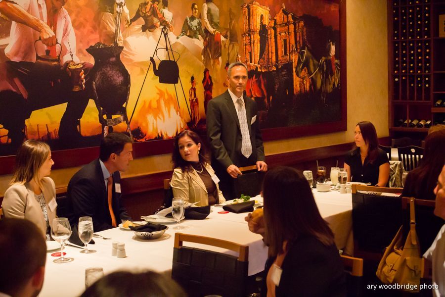 Man speaking at a table, surrounded by others in a restaurant; large mural on wall behind.