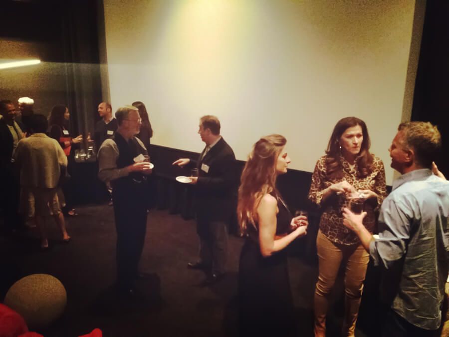 People socialize in a dark room near a large screen. Some hold drinks; the atmosphere is social.