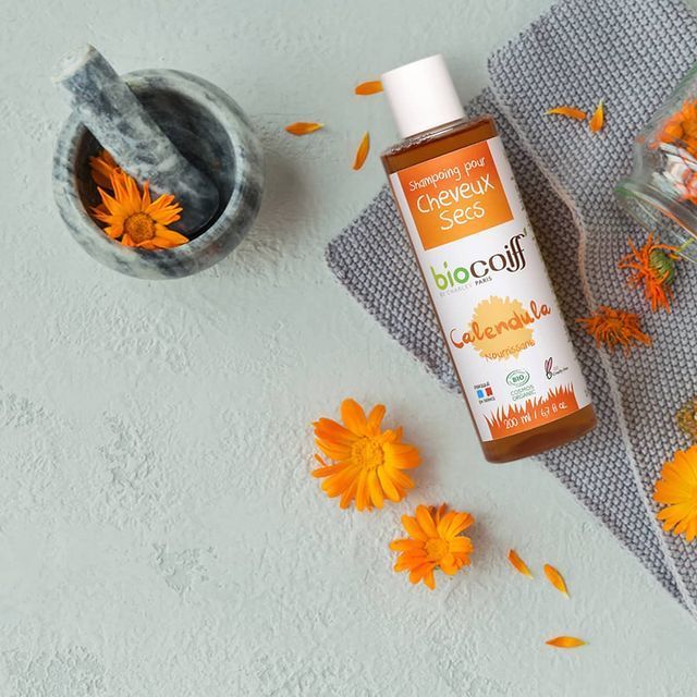 Oranje fles Bio-Oil met goudsbloemen naast een grijze vijzel en doek op een lichtgekleurde achtergrond.
