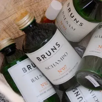 Diverse flesjes BRUNS haarverzorgingsproducten in een gevlochten mandje, met witte etiketten en gouden dopjes.