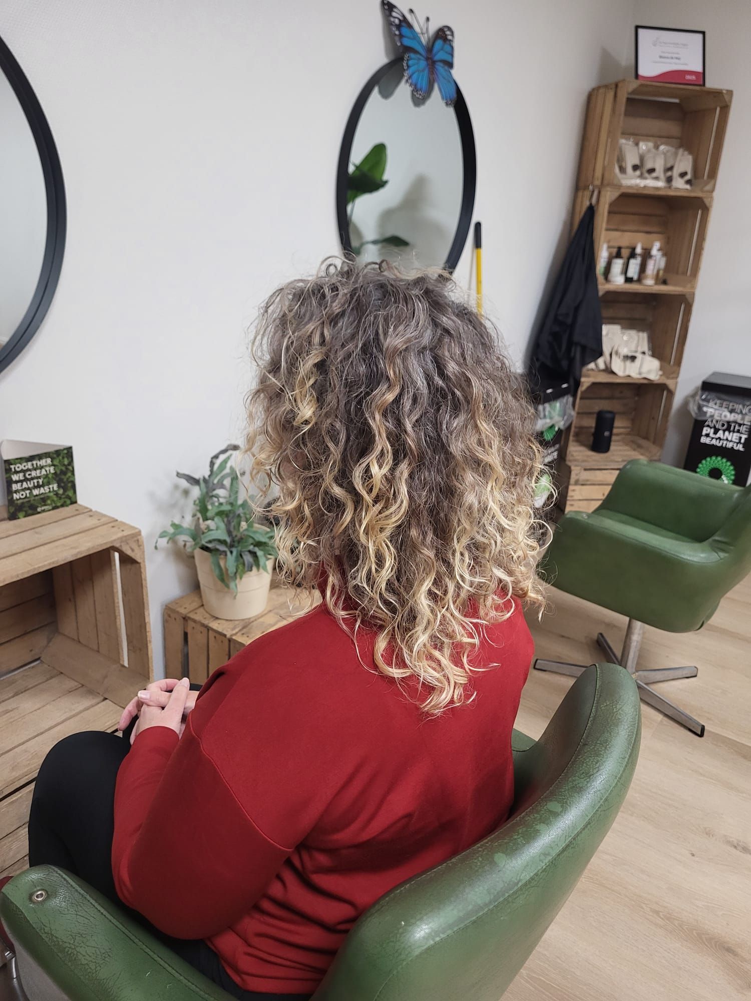 Een persoon met krullend haar en een rode trui zit in een groene stoel in een kapsalon, van achteren gezien.