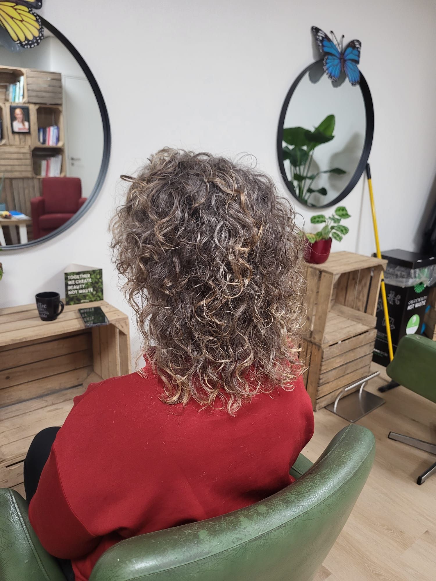 Een persoon met krullend haar en een rode trui zit in een groene stoel voor een spiegel in een gezellige kamer.