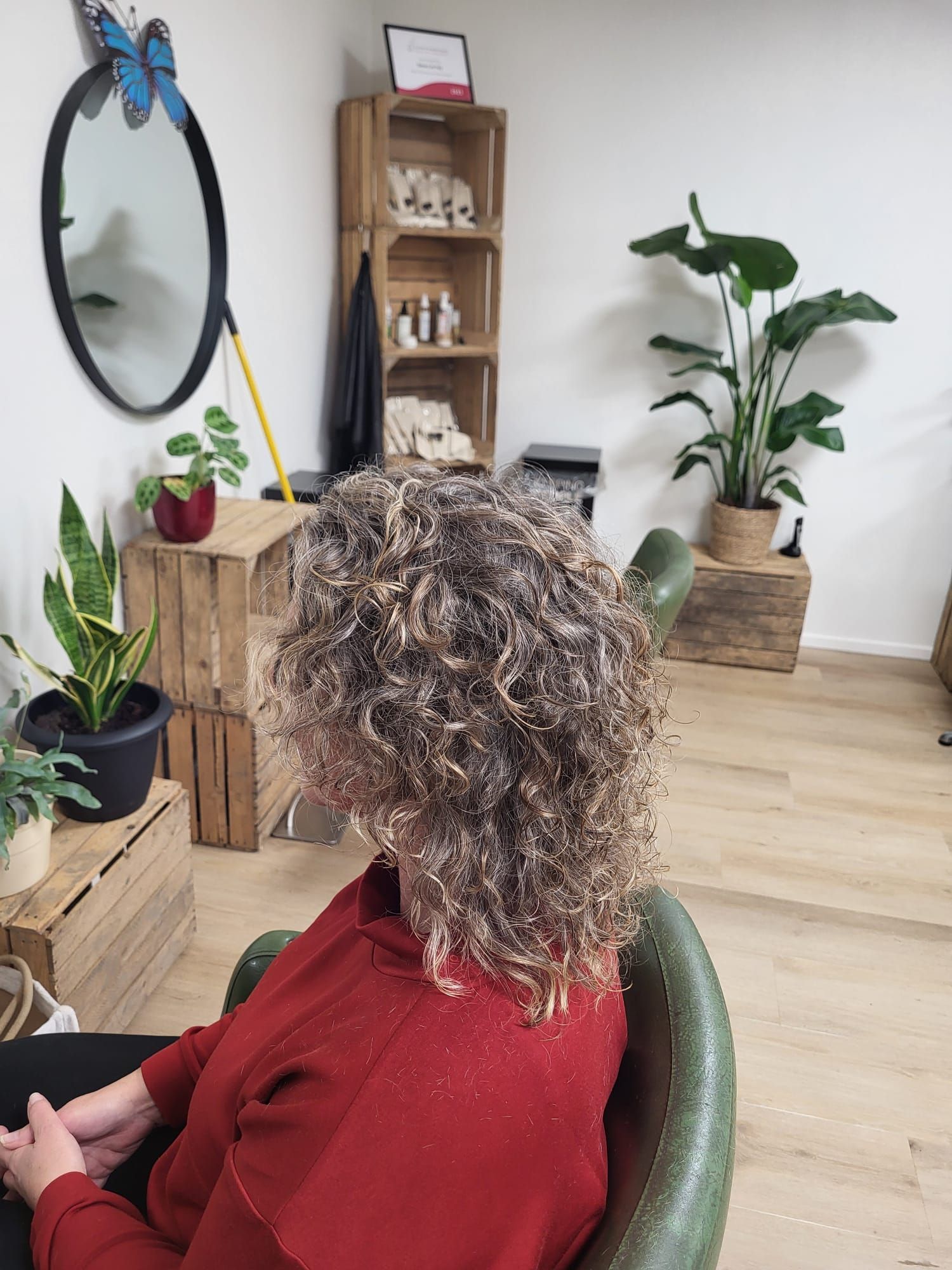 Persoon met krullend haar in een rode trui zit in een lichte kamer met planten en een spiegel.