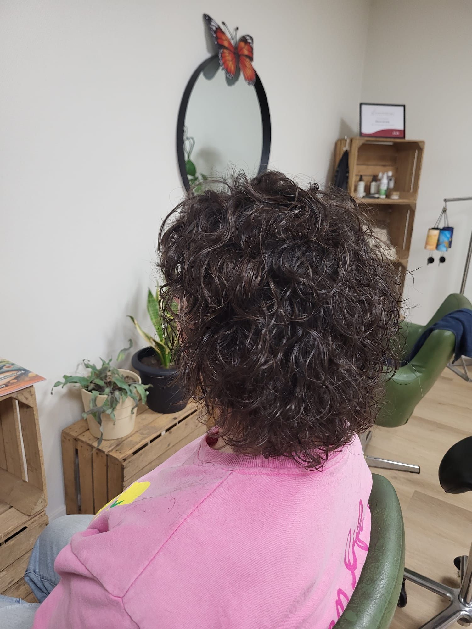 Een persoon met krullend donker haar zit in een groene stoel en kijkt in een spiegel in een gezellige kamer met planten en planken.