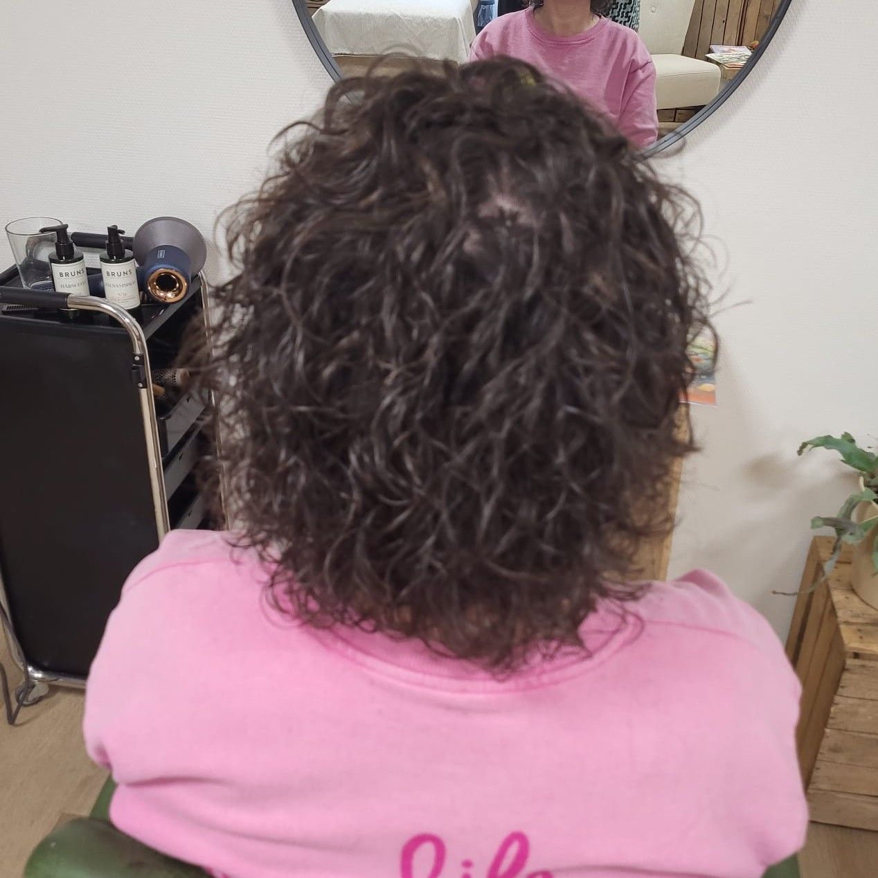 Achteraanzicht van een persoon met krullend haar in een roze shirt, die binnen bij een spiegel staat.