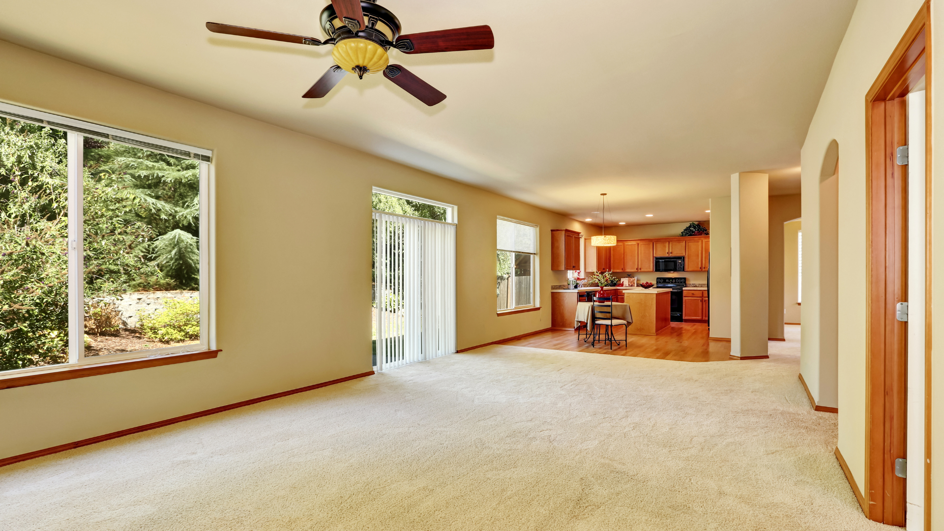 Spacious beige living room with ceiling fan, large windows, carpet, and kitchen area in back.