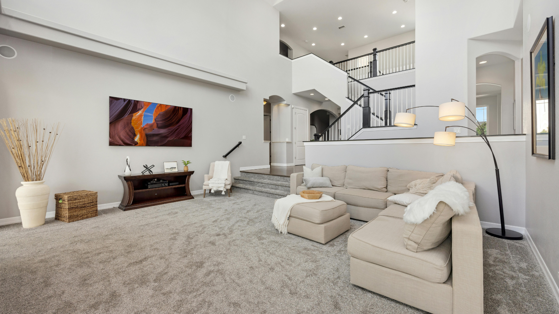 Bright modern living room with white sofas, gray carpet, artwork, and staircase