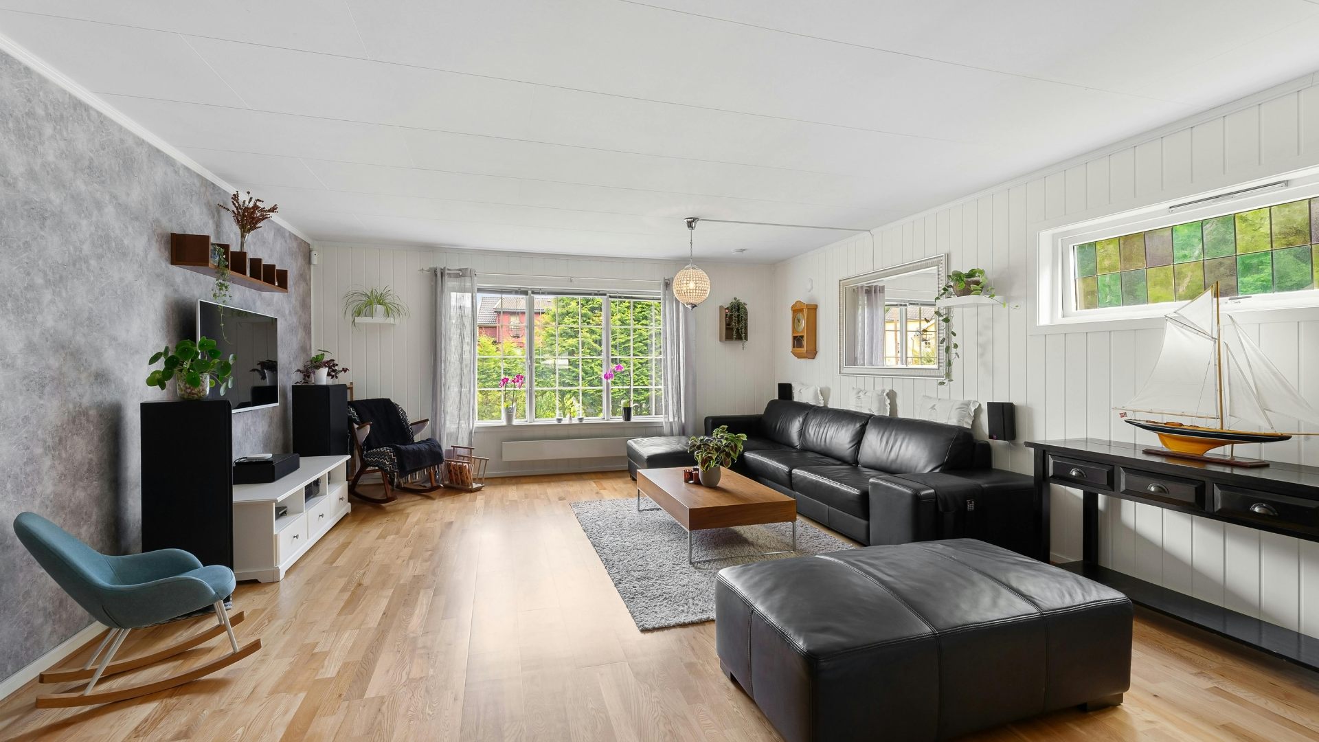 Bright modern living room with black sectional sofa, ottoman, TV stand, and large windows with greenery outside
